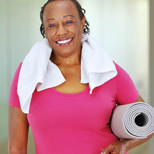 A smiling woman in a pink shirt with a white towel around her neck and a yoga mat rolled under her left arm