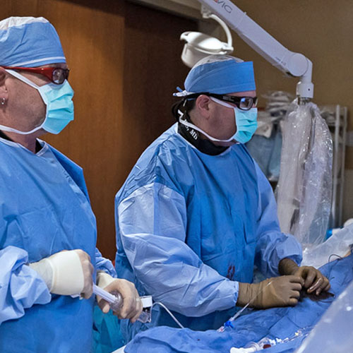 Two interventional cardiologists in blue scrubs perform a minimally invasive heart procedure in the operating room
