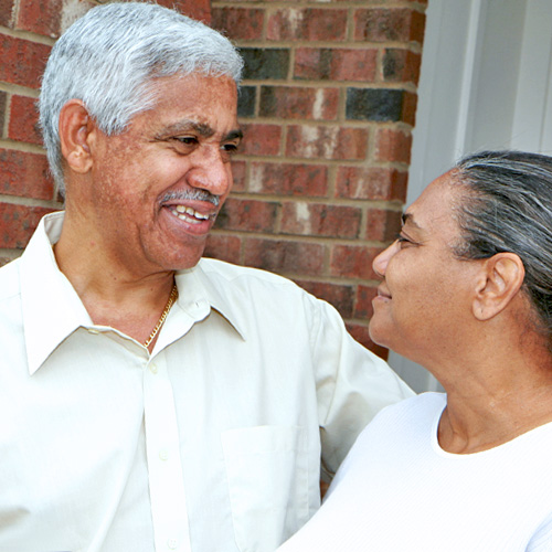 An older man in a light-colored button-down shirt smiles at a woman wearing a white shirt as they stand on a brick porch
