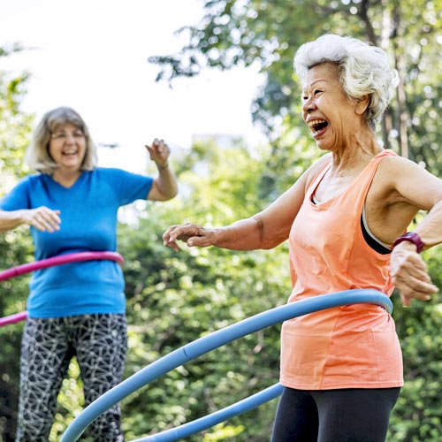 A woman in a blue T-shirt hula-hoops next to a hula-hooping older woman in an orange tank top in the park as part of their cardiac rehabilitation