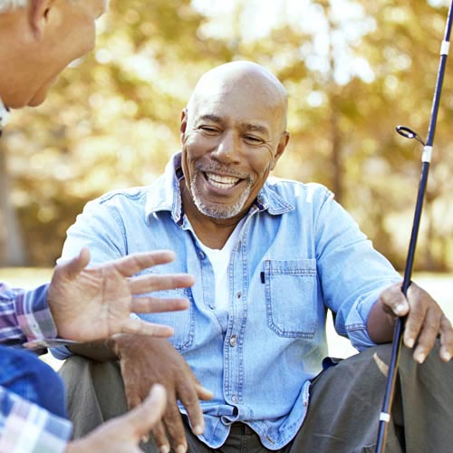 A man in a blue shirt sitting down holding a fishing pole talks with an older man