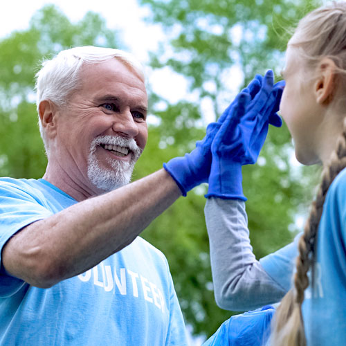 An older man wearing blue gloves and a blue shirt that says "volunteer" high-fives a young girl who is also wearing gloves and the same blue volunteer shirt